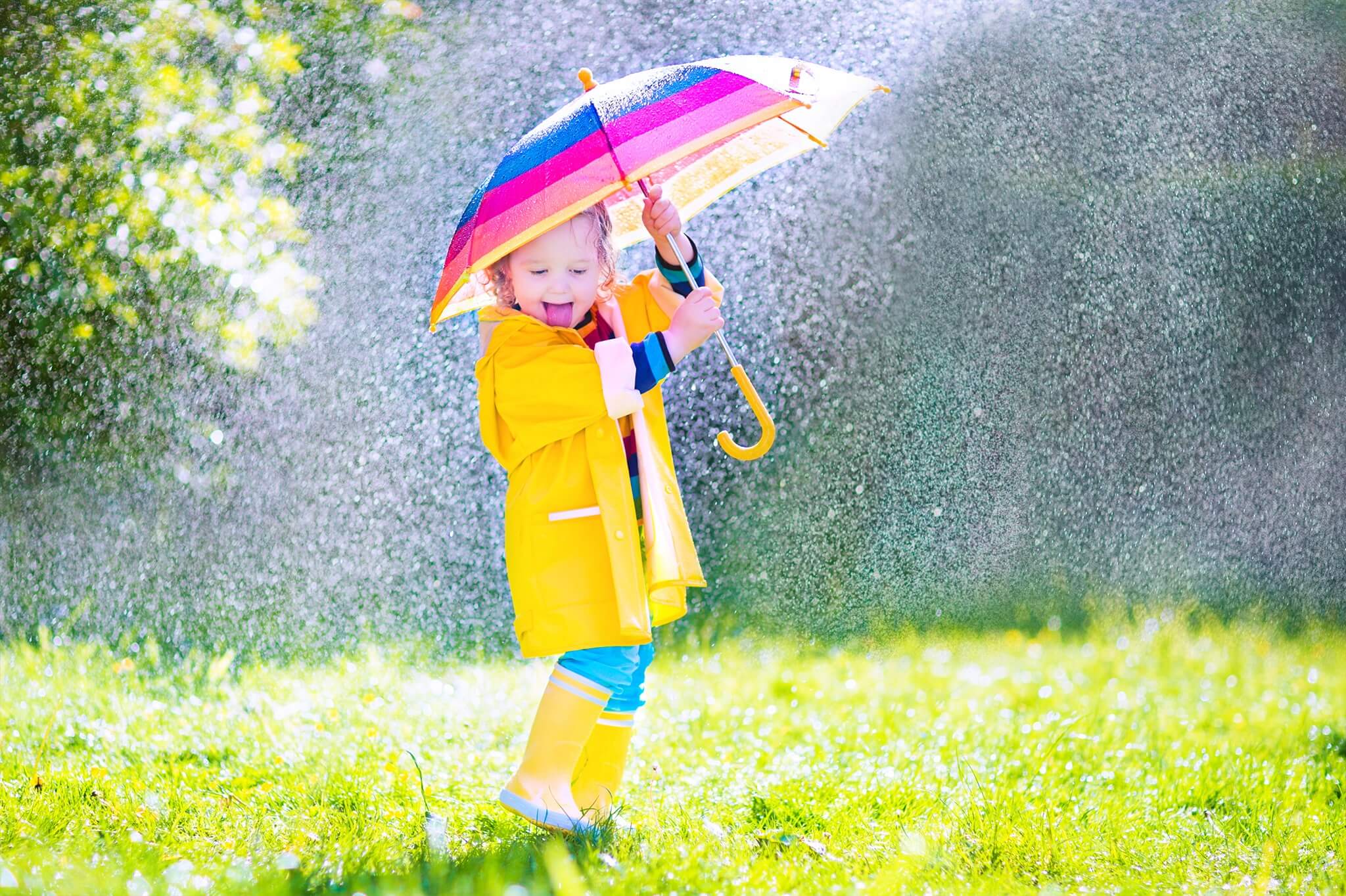 Kleinkind-Mädchen in gelben Regenklamotten und Gummistiefel freut sich unter einem buntegestreiften Regenschirm im Regen
